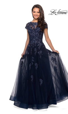 A woman elegantly holds the sides of the Long Formal Dress: La Femme 26907 in navy, a floor-length A-line gown featuring short sleeves, intricate floral embroidery, and embellished lace appliques.