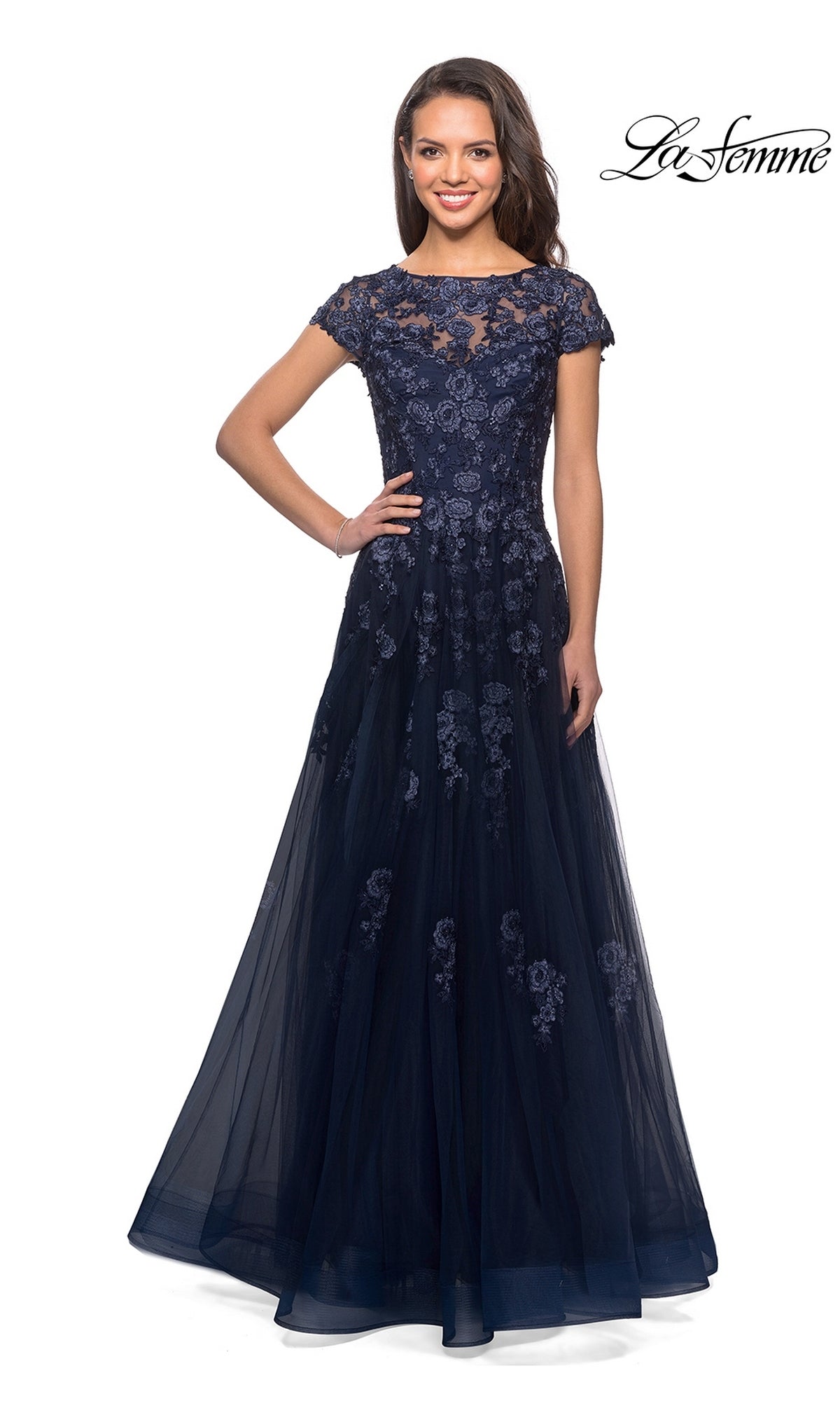 A woman wears the La Femme 26907, a floor-length, navy blue long formal dress featuring short sleeves, floral lace detailing, and a sheer overlay. Embellished lace appliques add texture to the elegant A-line silhouette. The La Femme logo is displayed in the top right corner.
