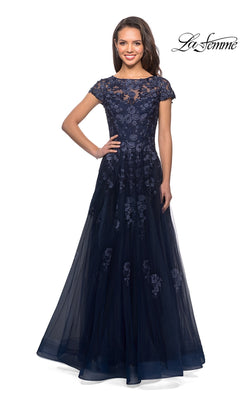A woman wears the La Femme 26907, a floor-length, navy blue long formal dress featuring short sleeves, floral lace detailing, and a sheer overlay. Embellished lace appliques add texture to the elegant A-line silhouette. The La Femme logo is displayed in the top right corner.