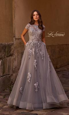A woman in the silver La Femme 26907 long formal dress, featuring an A-line silhouette with floral embroidery and embellished lace appliques, stands on a cobblestone street near a stone wall.