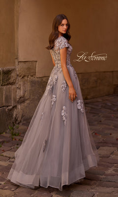 A woman wearing the La Femme 26907 long formal dress—a floor-length, lavender tulle gown with an A-line silhouette and delicate embellished lace appliques—stands on a cobblestone path, gazing over her shoulder. The “La Femme” logo appears in the image.