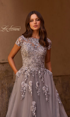 A woman stands against a neutral background, looking at the camera with her hands in the pockets of a gray La Femme 26907 long formal dress. The gown features floral embroidery and embellished lace appliques in an elegant A-line silhouette. “La Femme” is written in script on the left.