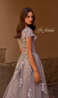 A woman stands sideways, looking over her shoulder against a neutral background, wearing the La Femme 26907 long formal dress—a sheer gown adorned with floral embroidery and embellished lace appliques. The “La Femme” logo appears near her head.