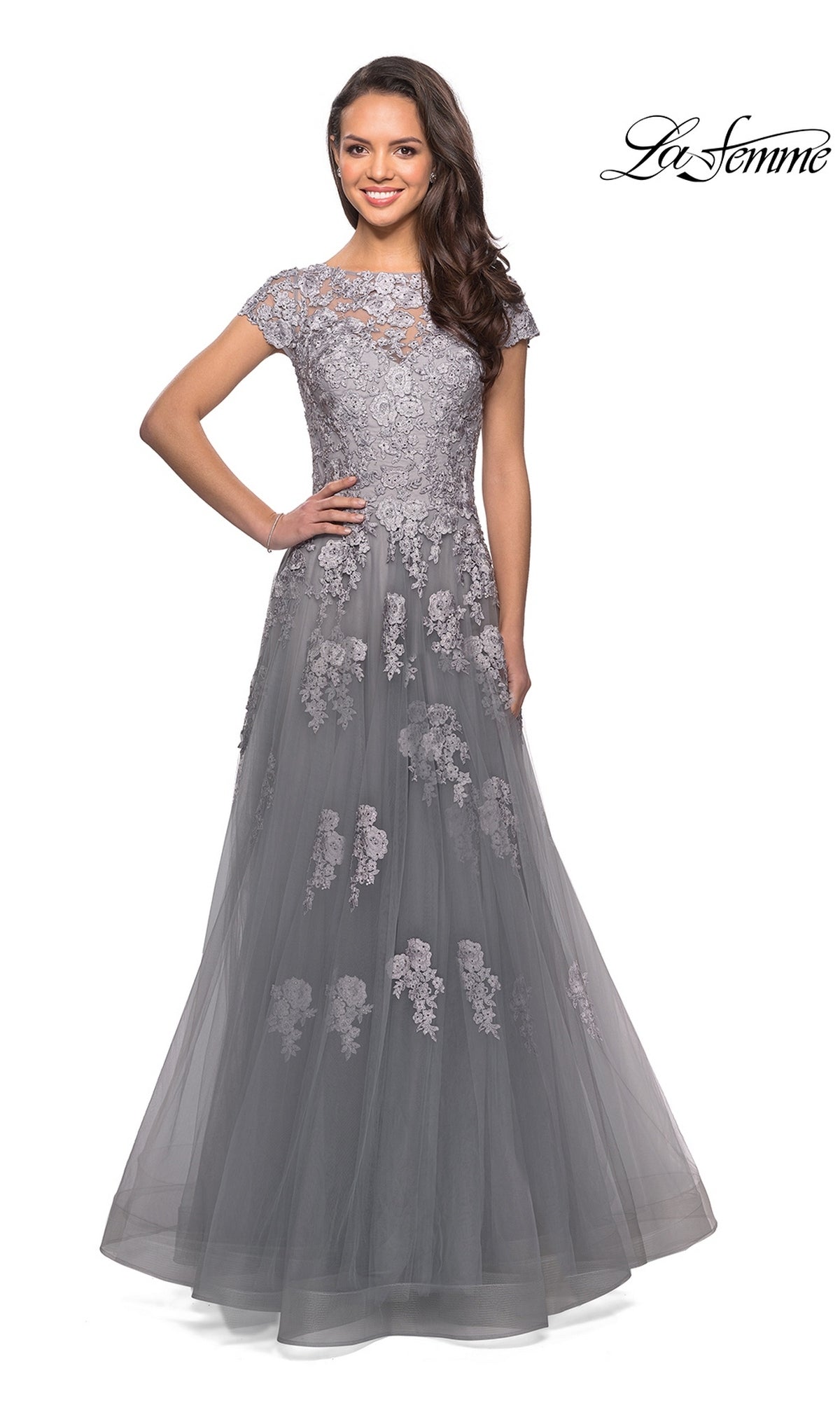A woman stands smiling with one hand on her hip, wearing the Long Formal Dress: La Femme 26907—a floor-length, short-sleeve gray gown featuring an A-line silhouette with embellished lace appliques. The “La Femme” logo appears in the top right corner.