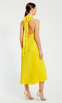 A woman faces away, wearing the Mac Duggal Tea Length Formal Dress 26909—sleeveless, pleated, tea length with a high neck and bow—paired with metallic high-heeled sandals.