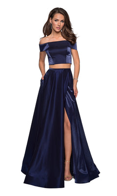A woman poses against a white background wearing the La Femme A-Line Two-Piece Satin Prom Dress 26919, featuring a navy blue off-the-shoulder crop top and matching high-waisted, floor-length skirt with an A-line silhouette and thigh-high slit.