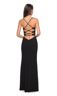 A woman stands facing away from the camera in the La Femme Strappy Open-Back Jersey Prom Dress 26940, a long black gown with crisscross straps and an open back.