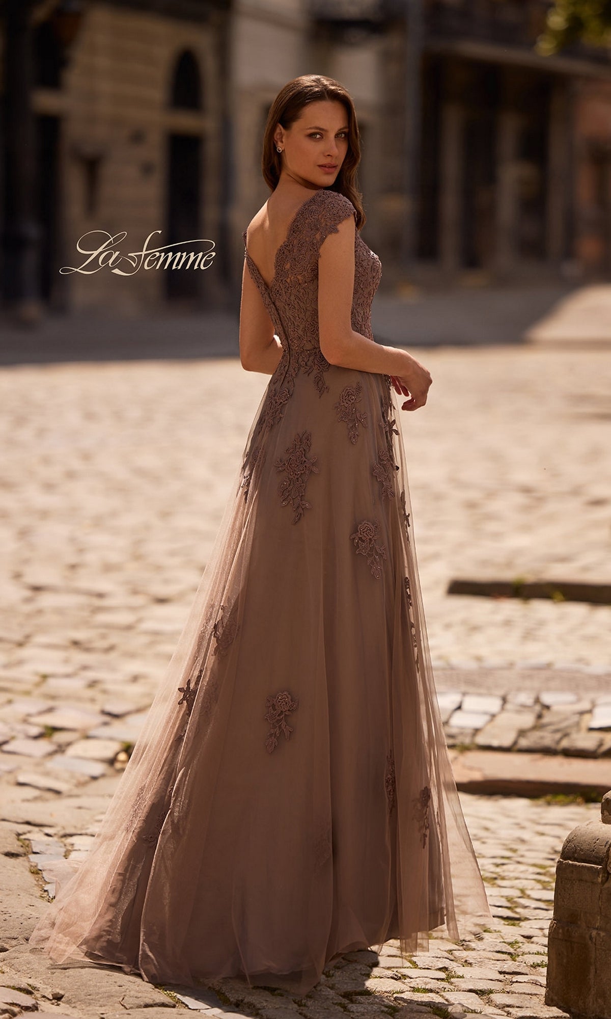 Woman stands on a cobblestone street wearing the La Femme 26942 Long Formal Dress—a floor-length, mauve gown with intricate floral lace details and cap sleeves, looking over her shoulder—the perfect elegant choice for any formal occasion.