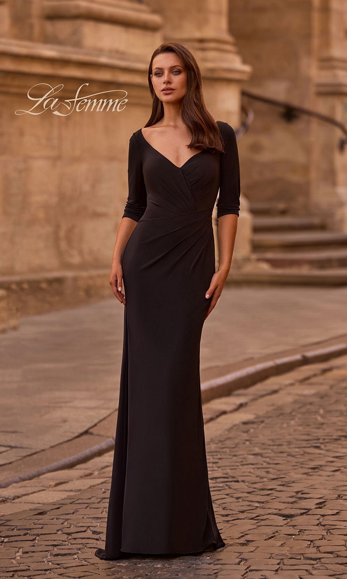 A woman stands on a cobblestone street in front of a historic stone building, wearing the La Femme 26955 long formal dress in black, featuring a floor-length fitted sheath skirt and three-quarter length sleeves.