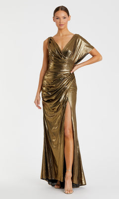 A woman in the Mac Duggal 26986 Long Metallic Gold Formal Dress with draped fabric and a thigh-high slit, paired with strappy heels, stands against a plain white background.
