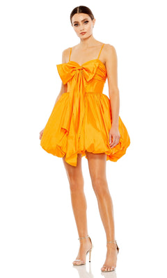 A woman wears the Short Party Dress 27040 by Mac Duggal, featuring a bright orange hue, large bow on the bodice, playful balloon hem, and styled with silver high-heeled sandals.