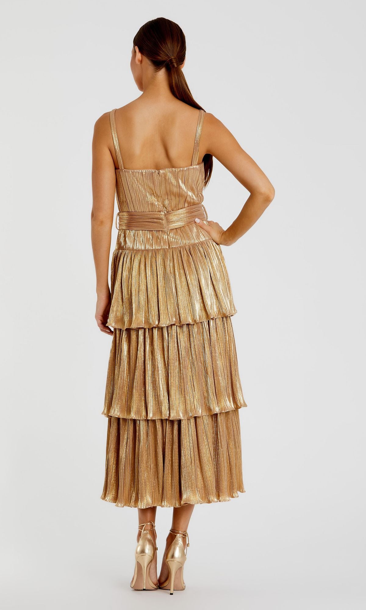 A woman faces away, wearing the Mac Duggal Tea-Length Metallic Party Dress 27062—a gold, pleated, tiered cocktail dress—paired with matching gold high heels.