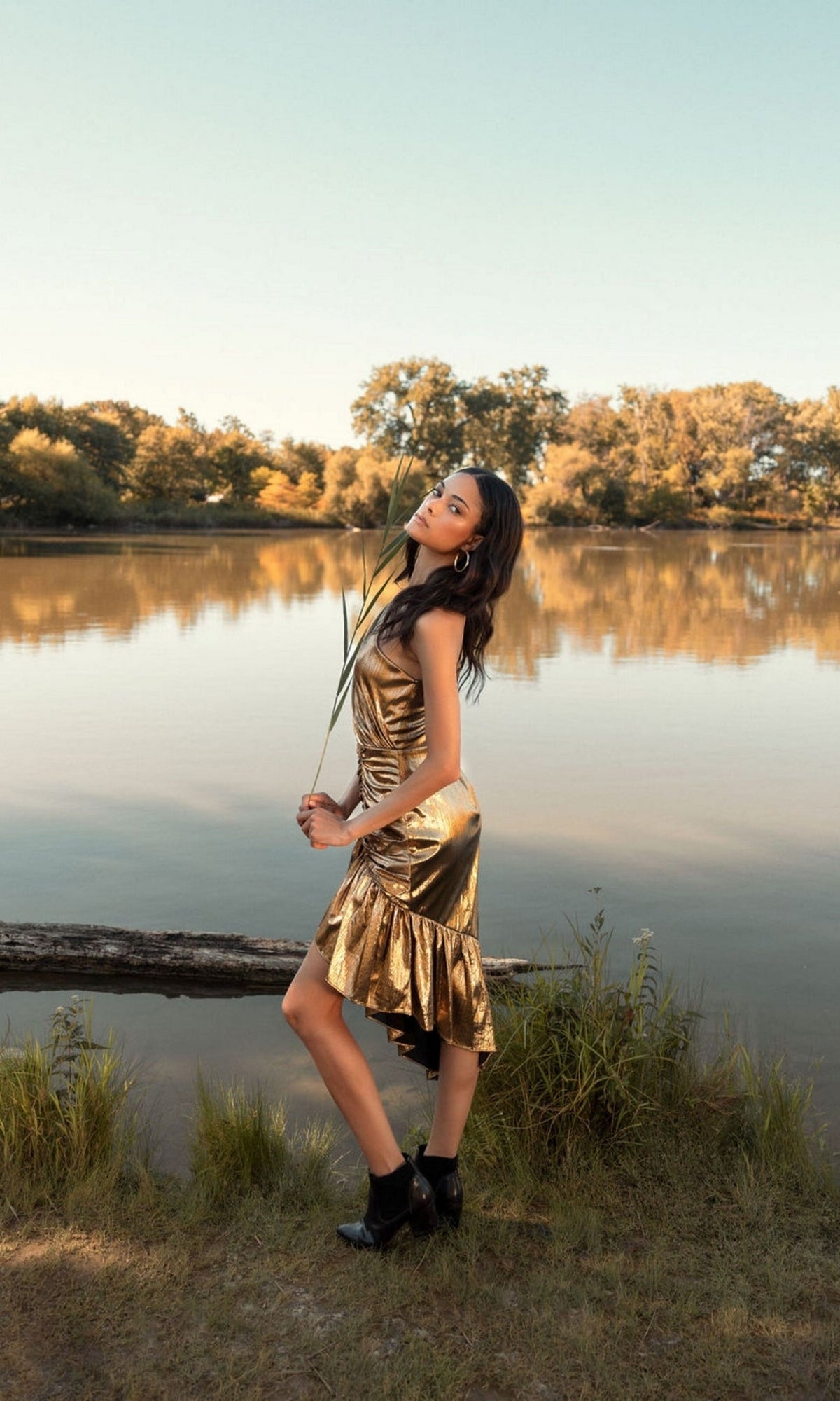 A woman in the Short Wedding Guest Dress 27075 by Mac Duggal and black boots poses by a calm lake, holding tall grass, with trees and blue sky in the background.