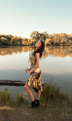 A woman in the Short Wedding Guest Dress 27075 by Mac Duggal and black boots poses by a calm lake, holding tall grass, with trees and blue sky in the background.