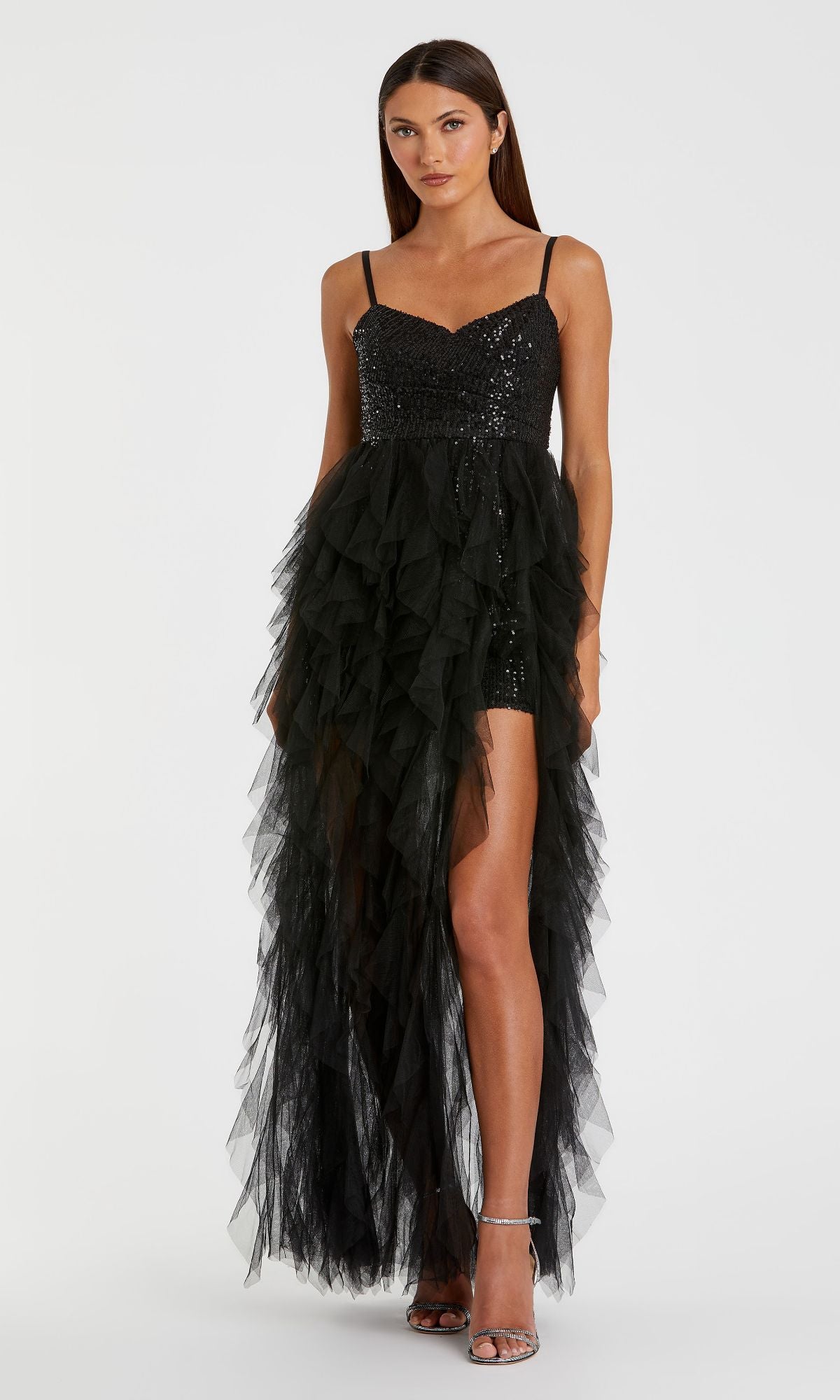 A woman wears the Mac Duggal 27119 Overskirt Mini Sequin Prom Dress—black, short, with a ruffled tulle skirt and high slit. Facing forward in silver heels, she flaunts the sparkling dress’s modern elegance.