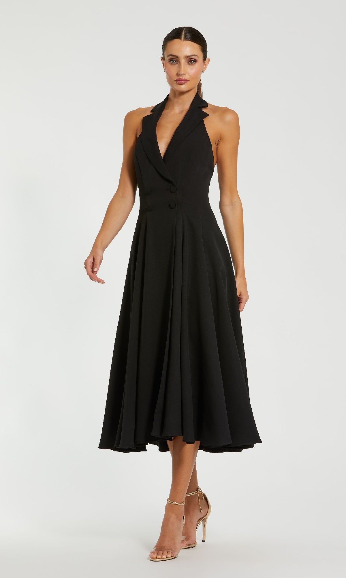 A woman models the Mac Duggal 27252 Blazer Halter Midi Party Dress, a sleeveless black dress with a fitted bodice and flared skirt, paired with strappy high-heeled sandals against a plain background.