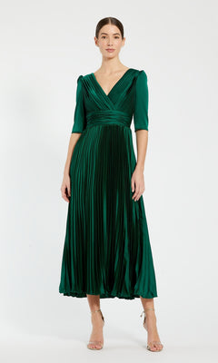A woman models the Long Formal Dress 27341 by Mac Duggal, featuring an emerald green, pleated A-line silhouette with elbow-length sleeves and a V-neckline, paired with nude heels against a plain background.