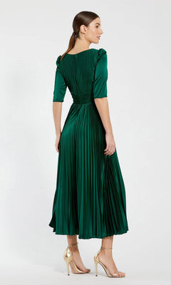A woman stands facing away, wearing the Long Formal Dress 27341 by Mac Duggal in emerald green—a pleated a-line midi dress with elbow-length sleeves—paired with gold high-heeled sandals.