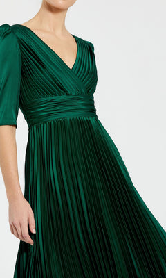 A woman stands against a plain background in the Long Formal Dress 27341 by Mac Duggal—an emerald green, pleated A-line gown with a V-neck, short sleeves, and a fitted waistband.