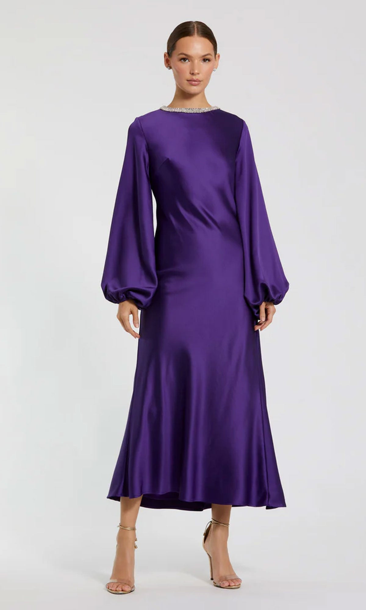 A woman wears the Mac Duggal 27350 Modest Midi Wedding Guest Dress—long sleeve, purple satin, round neckline, slight hem flare—paired with strappy nude heels.