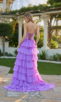 A woman stands outdoors on a stone path, facing away, wearing the Long Prom Dress: Juliet JT2738S, which features a strapless bodice and chiffon ruffle skirt with greenery and classical columns in the background.