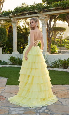 A woman stands outdoors in a garden, wearing the Juliet JT2738S long prom dress—a yellow tiered gown with a corset bodice and lace-up back—looking over her shoulder.