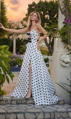 A woman in the Juliet JT2740S long prom dress in polka dot stands outdoors by a stone pillar, surrounded by greenery and flowers at sunset, wearing a strapless sweetheart neckline style with a thigh-high slit.