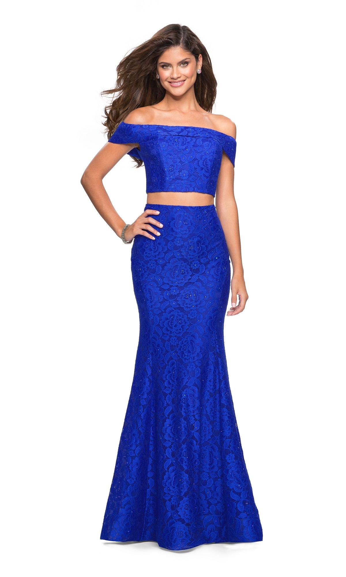 A woman smiles in the La Femme Two-Piece Mermaid Lace Prom Dress 27443, featuring royal blue off-the-shoulder straps and a fitted floral lace top, standing against a plain white background.