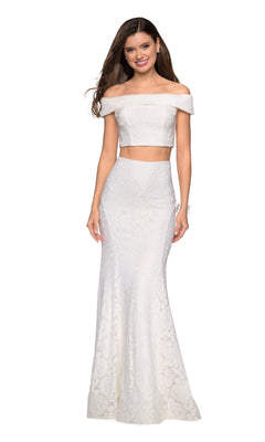 A woman wearing the La Femme Two-Piece Mermaid Lace Prom Dress 27443—a white, off-the-shoulder lace gown with a fitted crop top and floor-length skirt—stands and smiles.
