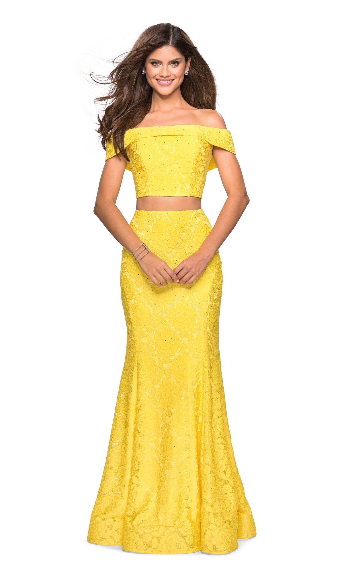A woman wears the La Femme Two-Piece Mermaid Lace Prom Dress 27443, a bright yellow off-shoulder gown with floral lace details, standing and smiling with her hands clasped in front.