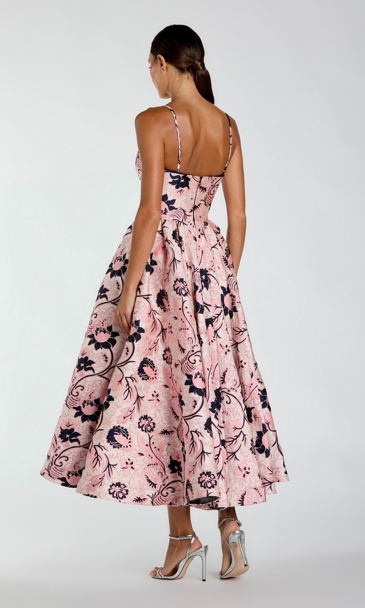 A woman stands facing away, wearing the Mac Duggal 27473 Midi Party Dress—a sleeveless, pink floral party dress with a full-skirted midi silhouette—paired with silver high-heeled sandals against a plain background.