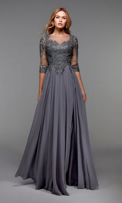 A woman wears the Alyce 27474 long formal dress—an elegant grey mother-of-the-bride gown featuring an embellished bodice and lace sleeves—standing against a plain grey background.