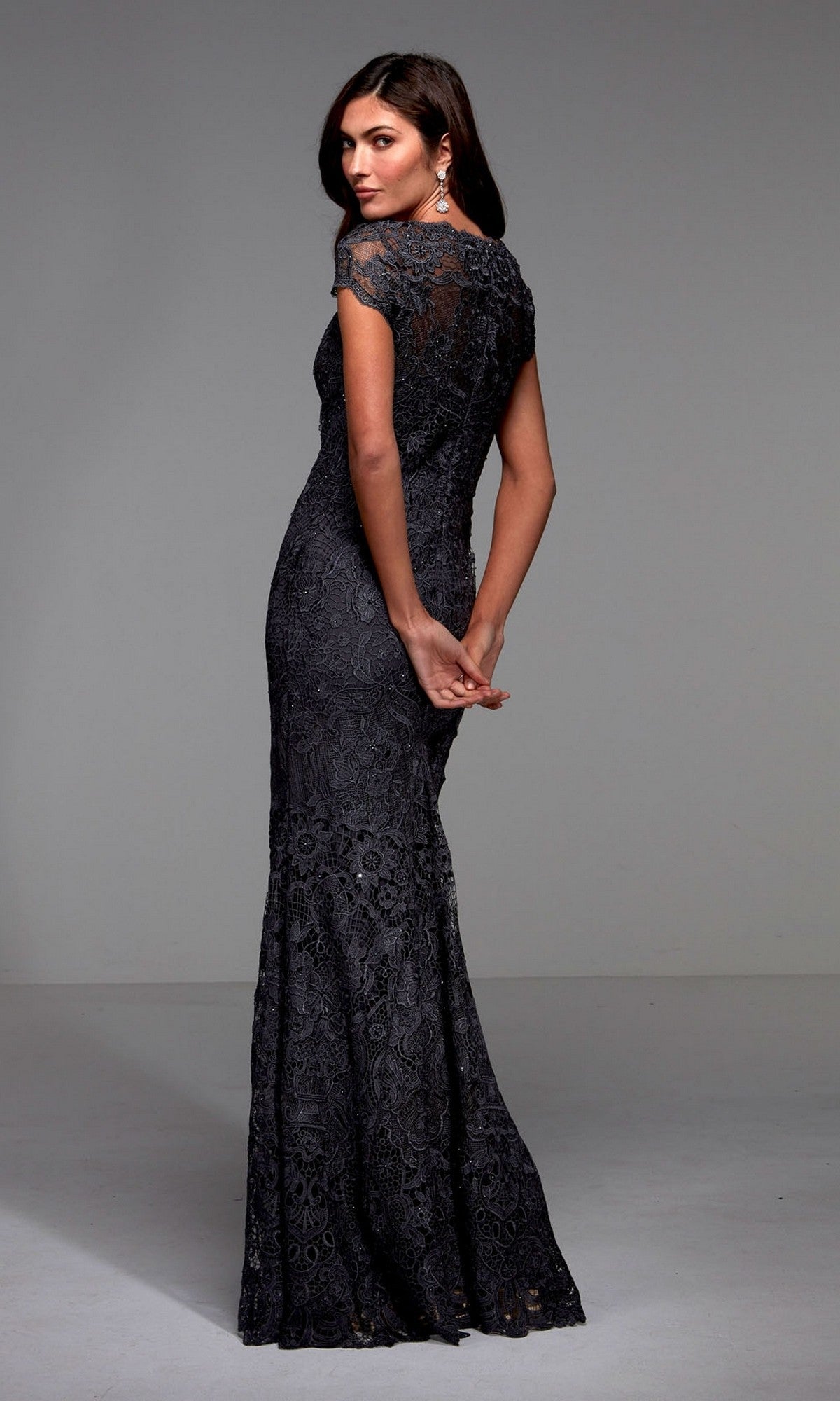 A woman stands with hands behind her back, facing away, wearing the Alyce 27478 floor-length formal dress in a dark color with short sleeves, lace details, and a v-neckline.