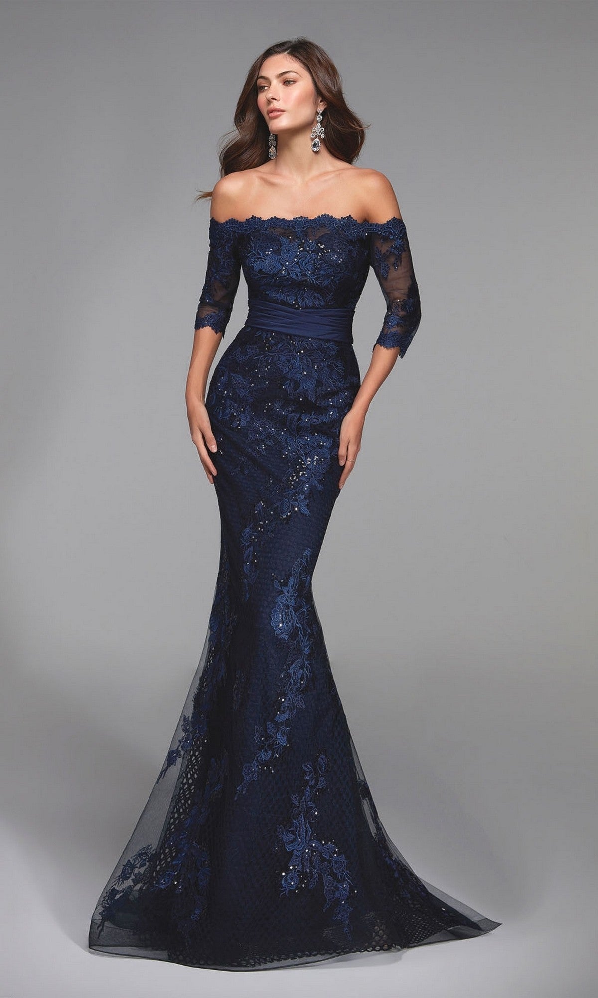 A woman poses against a plain gray backdrop in the Alyce 27495 long formal dress—a navy, off-the-shoulder, floor-length mermaid gown with beaded lace and sequin details, ideal for a mother-of-the-bride look.