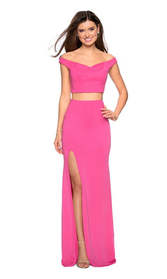 A woman smiles with one hand on her hip, wearing the La Femme 27496: a bright pink off-shoulder two-piece prom dress featuring a crop top and matching high-slit maxi skirt.