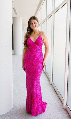 A woman stands indoors by large windows, wearing the Sleek Fitted Long Sequin Prom Dress by Aleta—a floor-length, bright pink gown with thin straps and shimmering sequins.