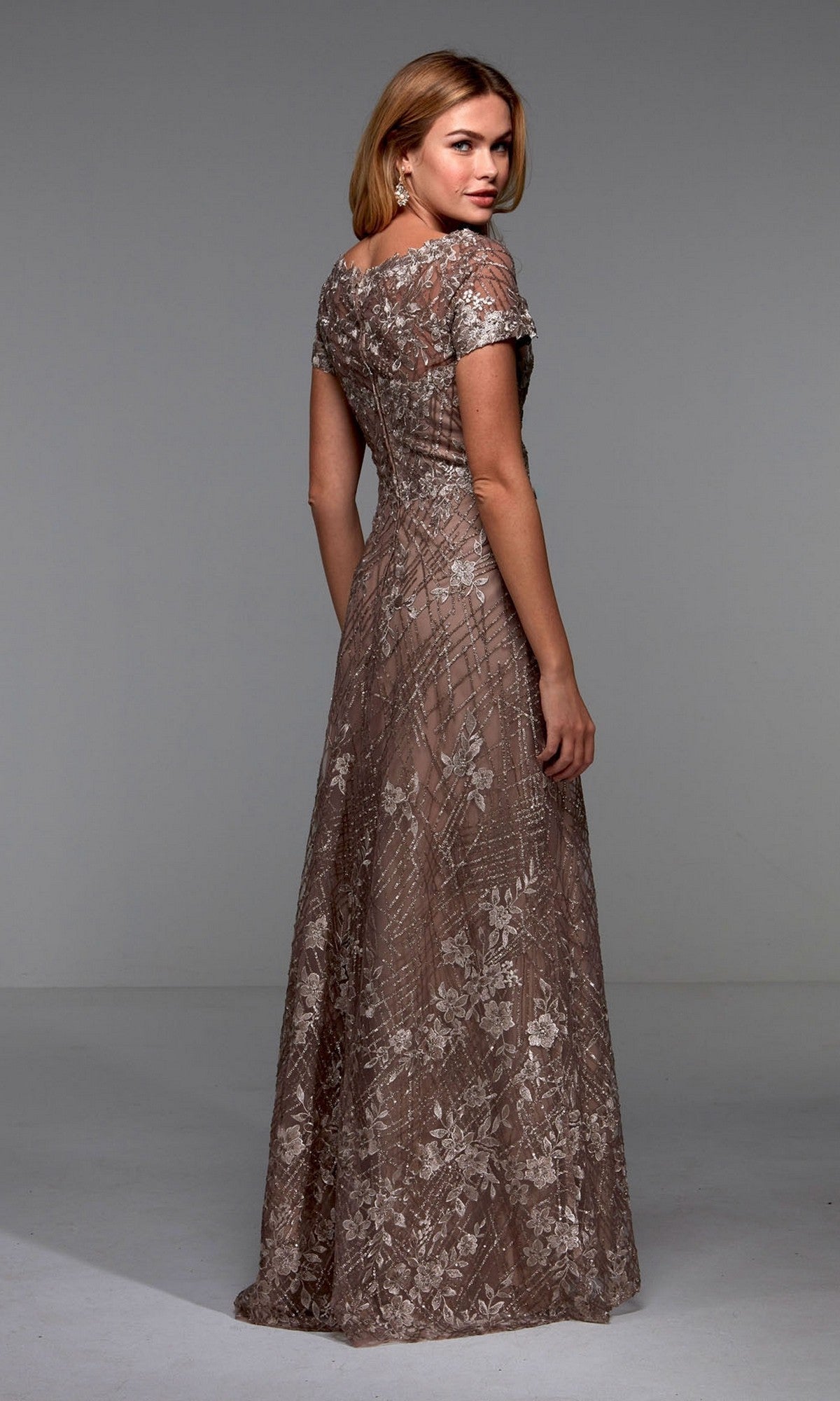 A woman faces away, wearing the Alyce 27525 long formal dress—a floor-length brown gown with short sleeves and floral lace appliques—posed in a simple studio setting.