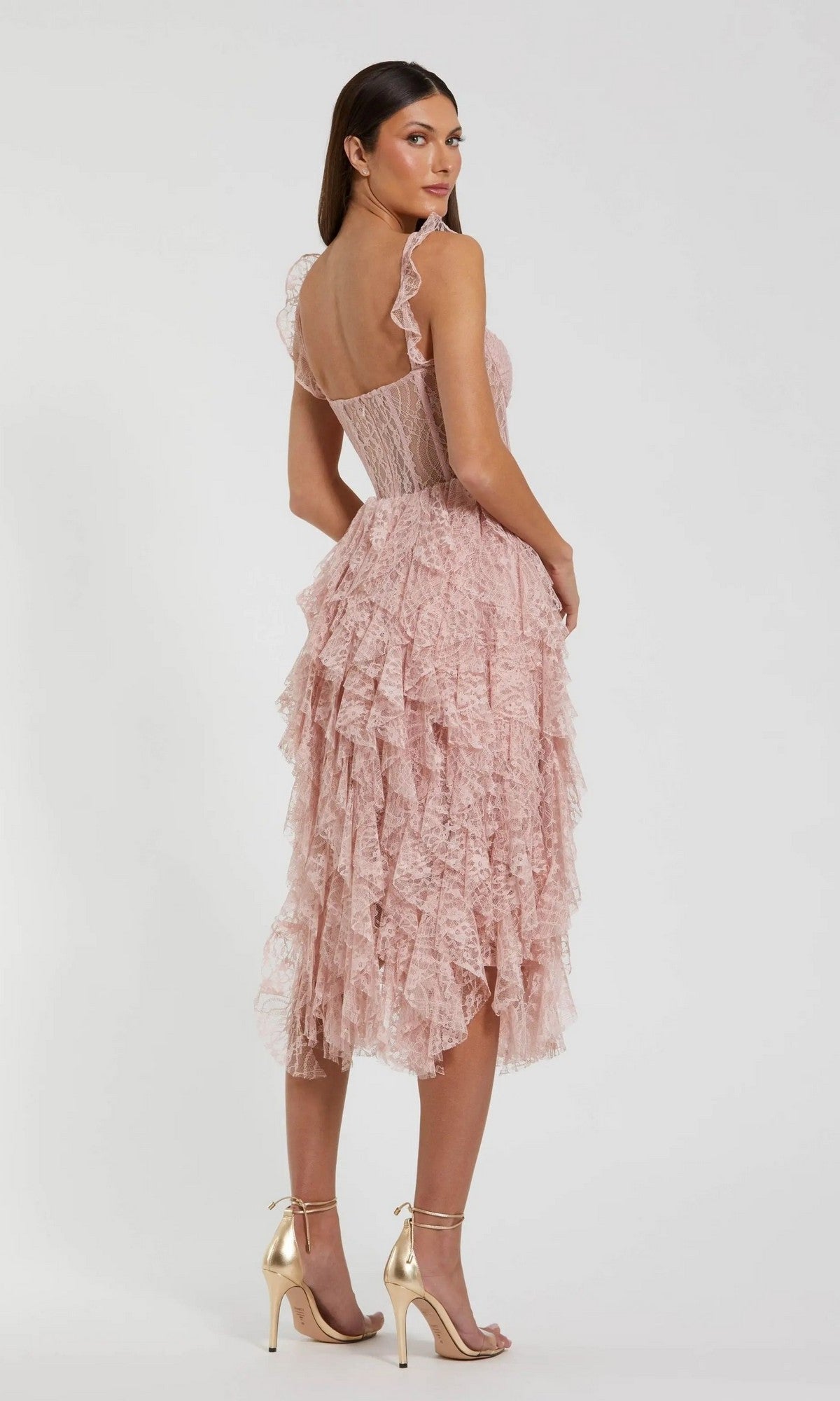 A woman stands facing backward with her head turned toward the camera, wearing the Mac Duggal 27529 Midi Party Dress—a light pink dress featuring a ruffled corset bodice, delicate lace detailing, sheer accents, and paired with gold high-heeled sandals.