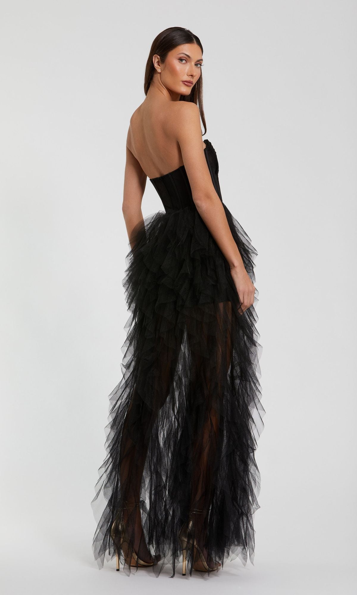A woman poses against a plain light background wearing the Mac Duggal 27532 Strapless Ruffled Long Prom Dress, featuring a fitted bodice and layered black sheer tulle ruffled skirt.