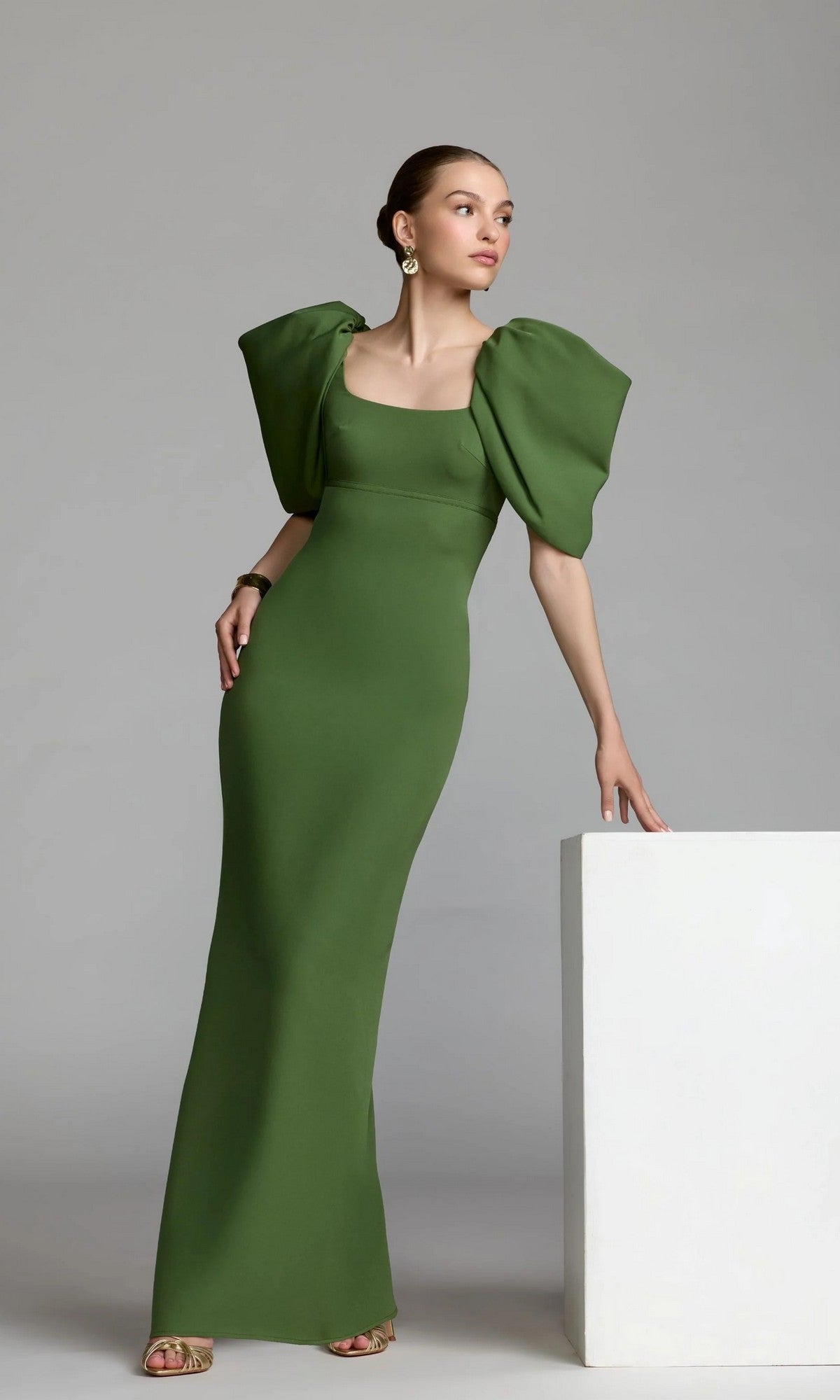 A woman in the Mac Duggal 27534 Puff-Sleeve Long Formal Dress, featuring a fitted green silhouette and large puffed sleeves, stands beside a white rectangular block against a plain gray background.