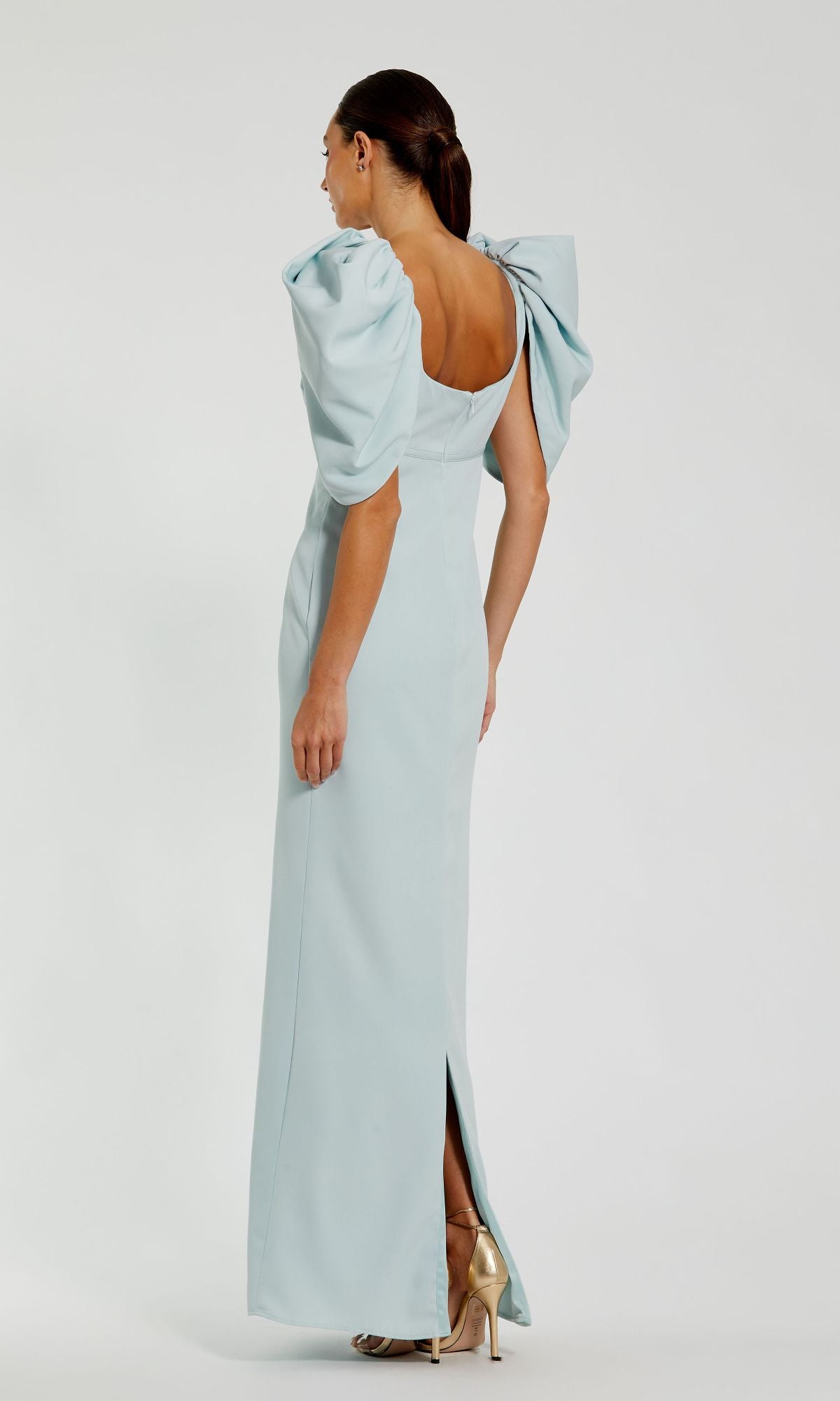 A woman faces away in the Mac Duggal 27534 Puff-Sleeve Long Formal Dress, featuring light blue fabric, puffed sleeves, and a back slit. Her long hair is pulled back and she wears gold high-heeled shoes to complete the look.