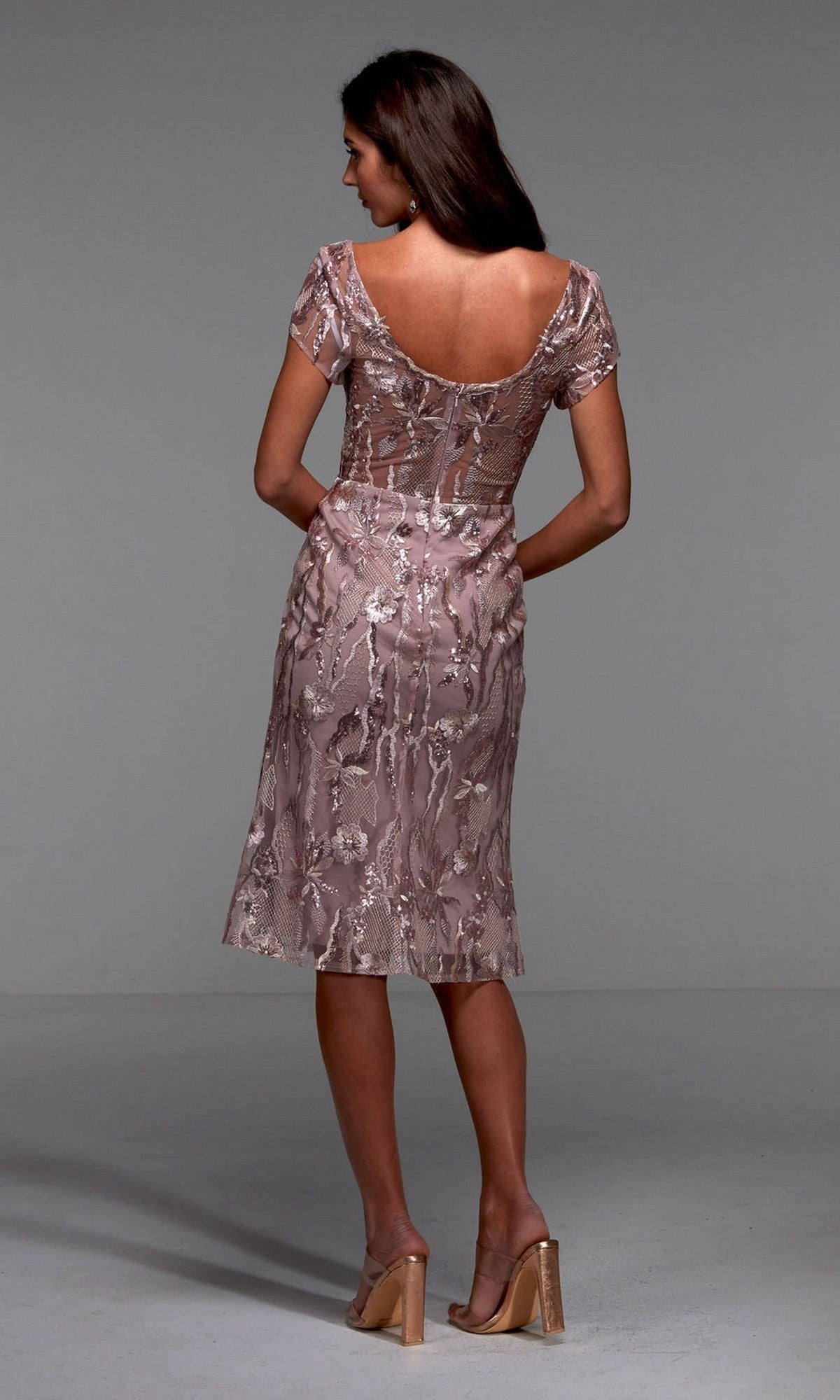 A woman stands facing away in the Alyce 27534 knee-length semi-formal dress—mauve with floral embroidery, sheer short sleeves, and a low back—paired with beige heeled sandals against a plain gray background.
