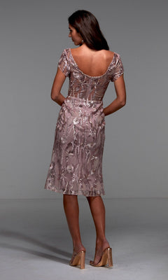 A woman stands facing away in the Alyce 27534 knee-length semi-formal dress—mauve with floral embroidery, sheer short sleeves, and a low back—paired with beige heeled sandals against a plain gray background.
