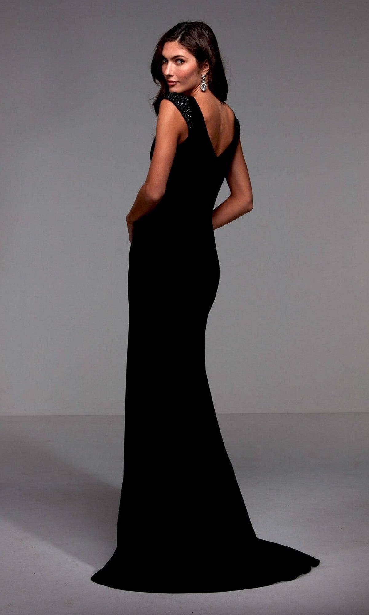 A woman wearing the Alyce 27539 long, black, sleeveless jersey formal dress stands facing away and looks over her shoulder, with visible earrings against a neutral background.