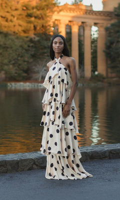 A person stands by a pond wearing a Mac Duggal 27540, a floor-length, cream-colored halter dress featuring large black polka dots and tiered ruffles.