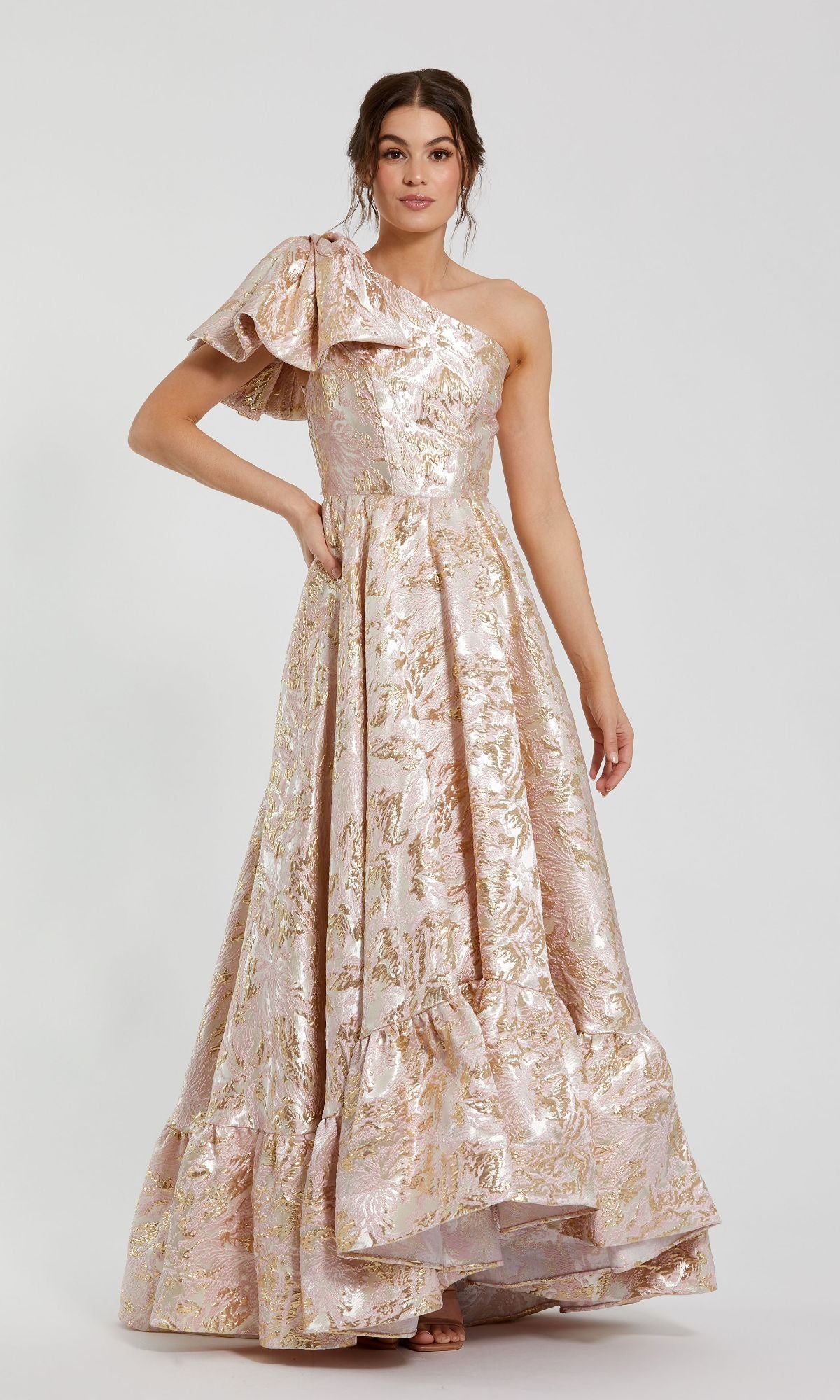 A woman in the Mac Duggal 27546 One-Shoulder Long Pink Ball Gown with ruffled sleeve and metallic floral pattern stands against a plain background.