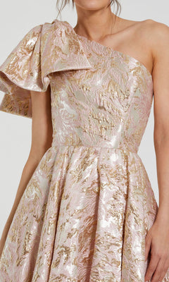 A woman wears the Mac Duggal 27546 one-shoulder long pink ball gown, featuring gold brocade fabric and a dramatic ruffle accent on the left shoulder.