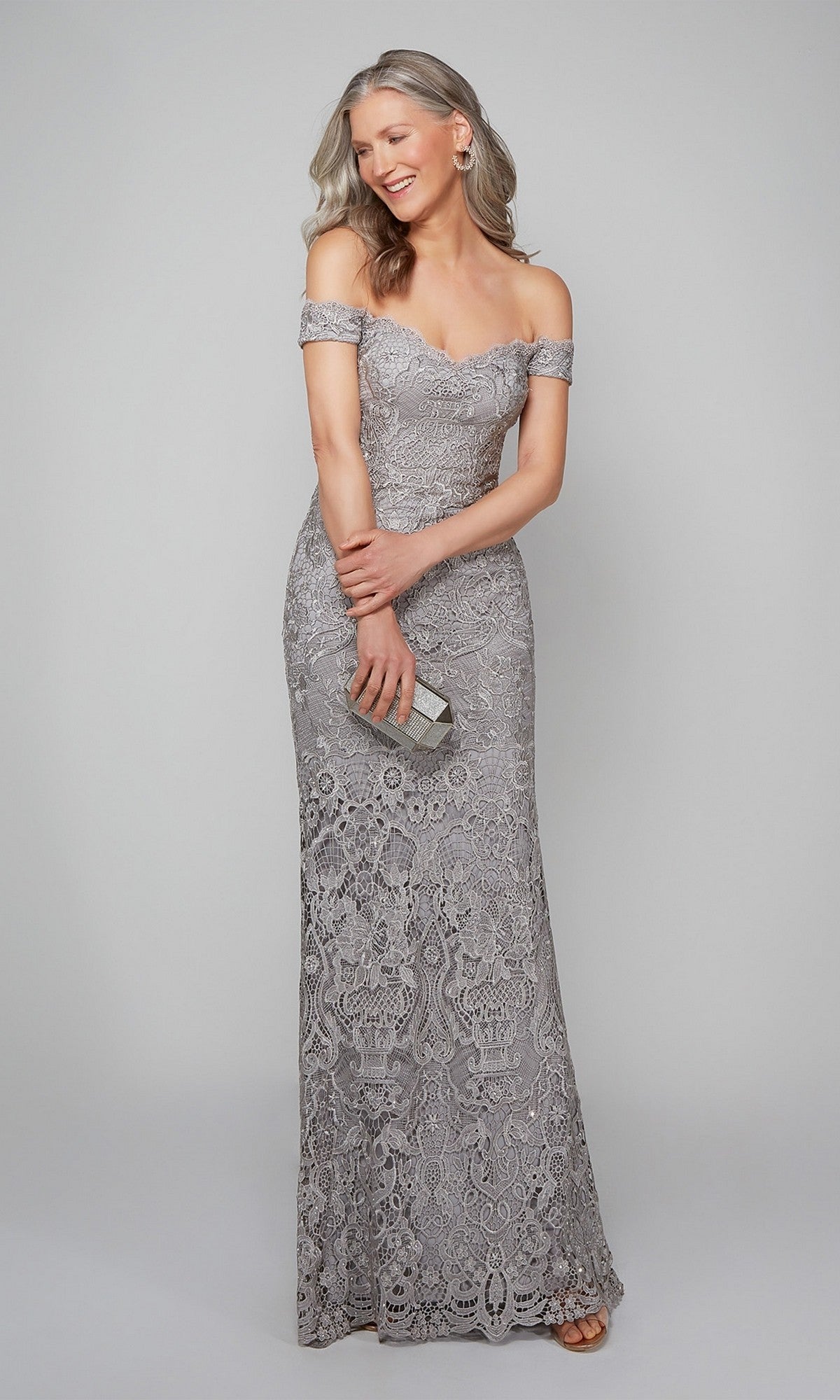 A woman in the Alyce 27553 long silver formal dress, featuring off-the-shoulder beaded lace, holds a small clutch and smiles against a plain light gray background.
