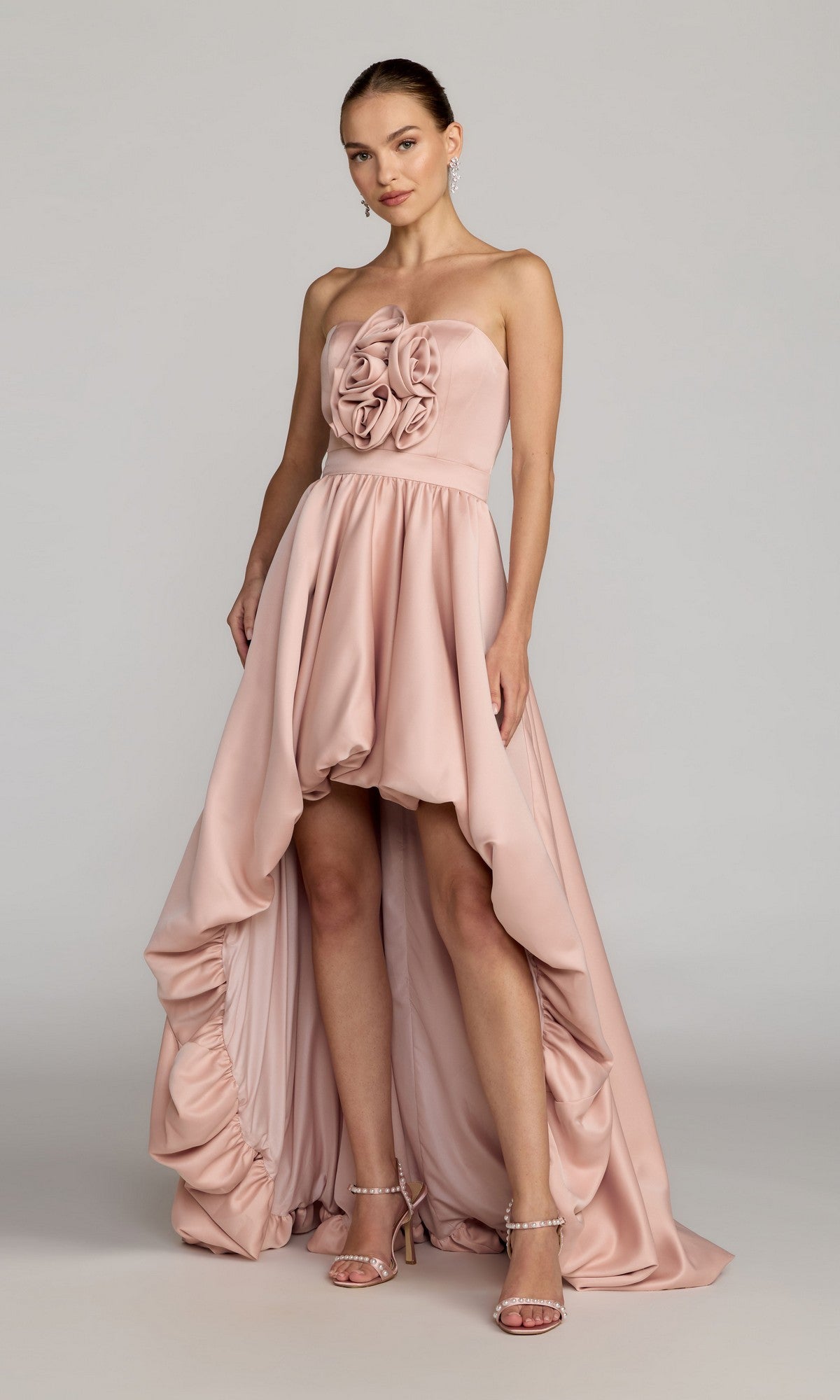 A woman wearing the Mac Duggal 27554 High-Low Formal Dress in blush, featuring a strapless bodice adorned with ruffled, rose-inspired details, elegantly paired with strappy heels.