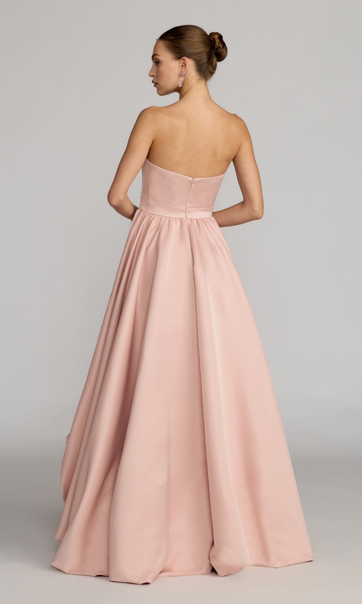 A woman stands with her back to the camera, hair styled in a bun, wearing the Mac Duggal 27554 High-Low Formal Dress—a strapless, floor-length blush pink gown that creates a timeless silhouette against a plain background.